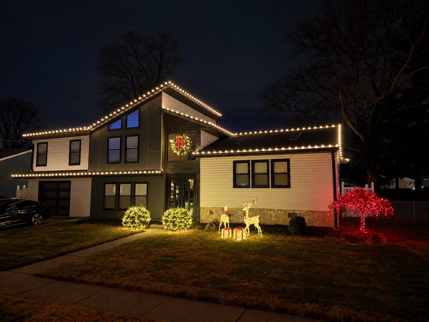 Split-level home at night with warm white roofline lights, a lit wreath over the entry, and light-up reindeer on the lawn