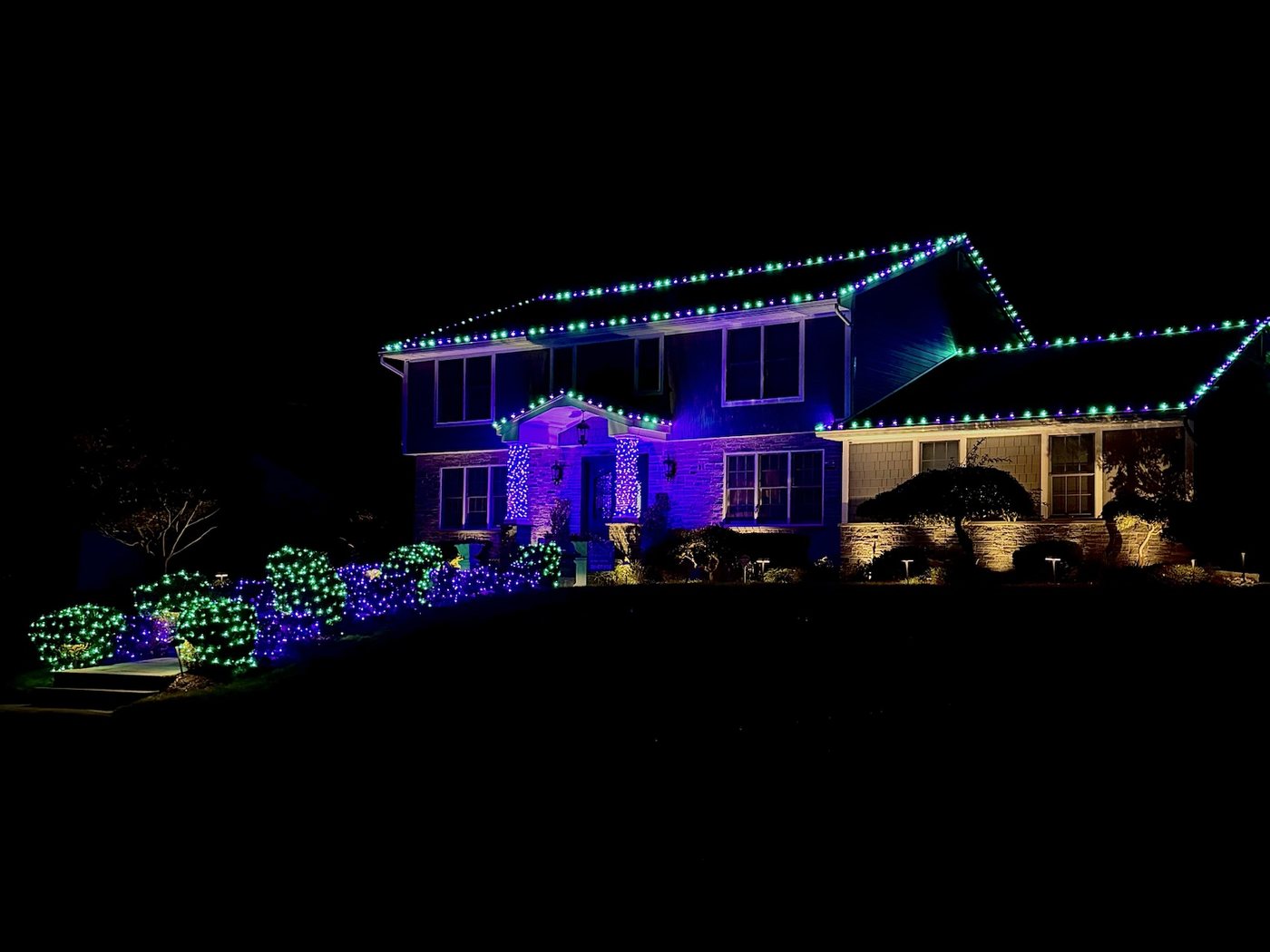 Two-story home with purple and green Halloween lights along the roofline and on the shrubs