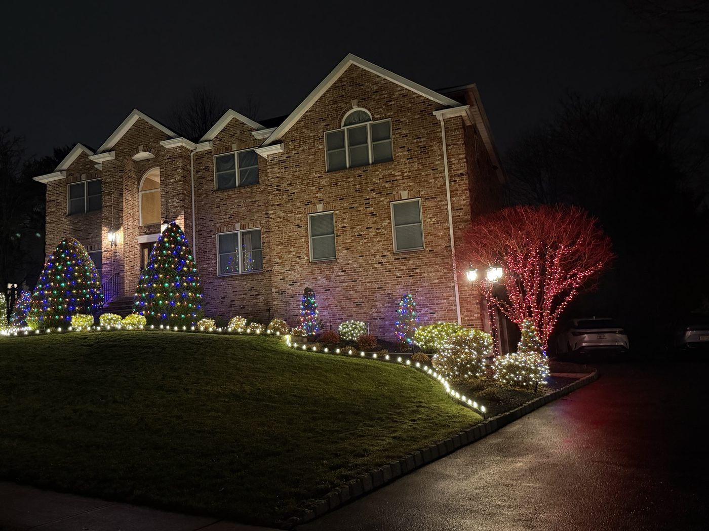 Brick colonial with multicolor and warm white lit shrubs, a red-lit tree, and a glowing walkway border