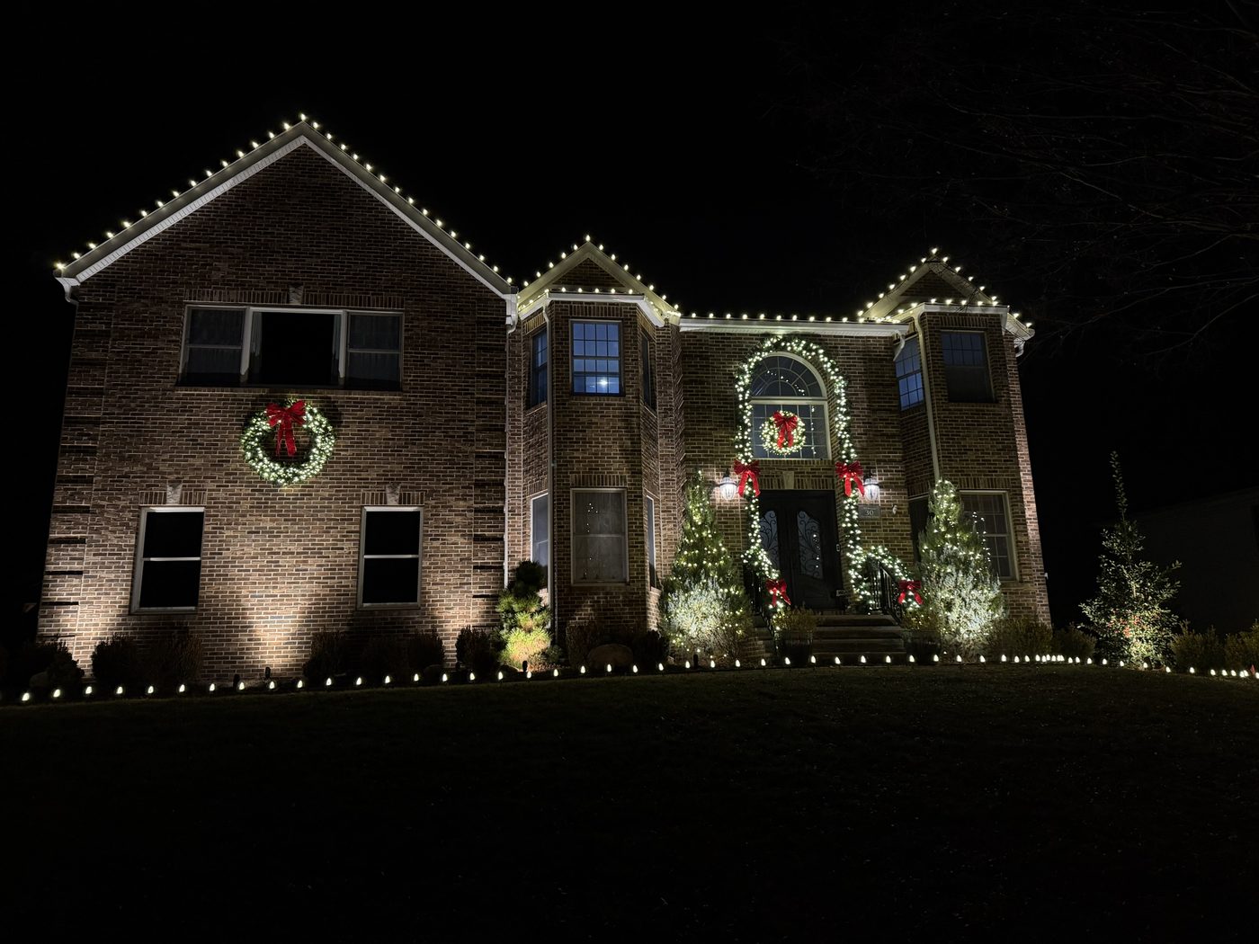 Brick colonial at night with warm white roofline lights, wreaths with red bows, and garland around the front entry